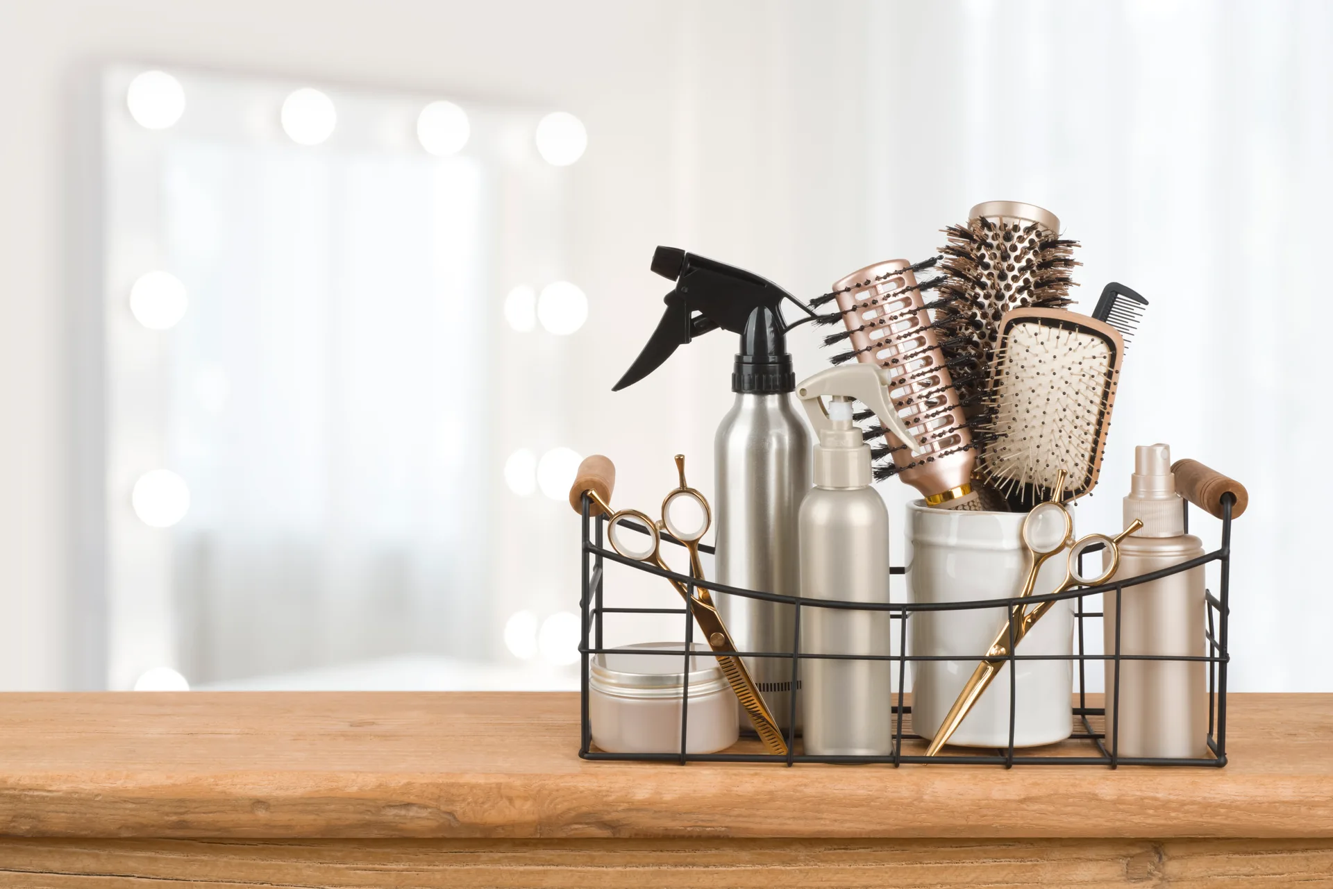 Wire basket of working tools on a wooden counter — gold shears, round brushes, spray bottles and combs against soft vanity light