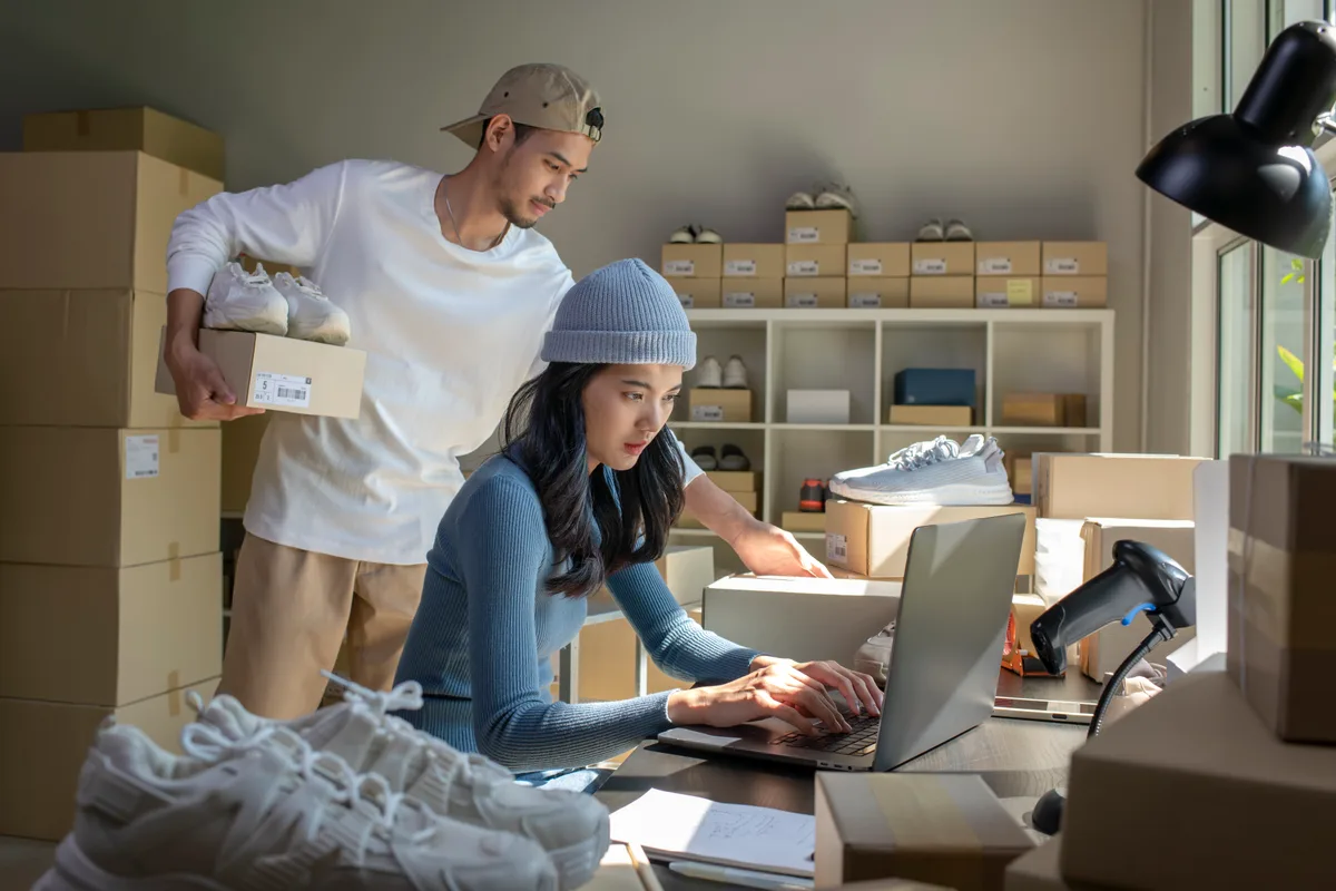 An online store owner packing customer orders at a laptop surrounded by shipping boxes