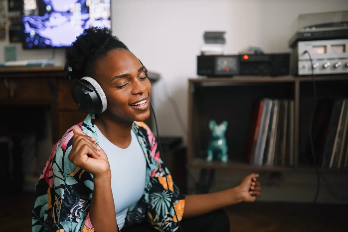 A listener smiling with eyes closed wearing wired over-ear headphones in a living room, with a turntable and record shelf behind her