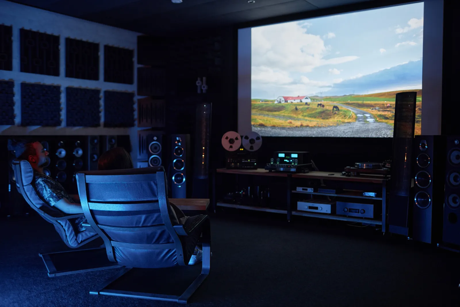The Drift Audio listening den at dusk: tower speakers, a reel-to-reel deck, and two listening chairs facing a projection screen