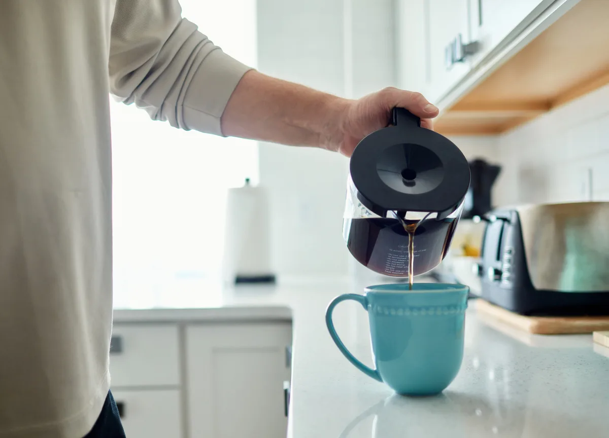 Person pouring freshly brewed coffee from a carafe into a blue mug in a sunny kitchen