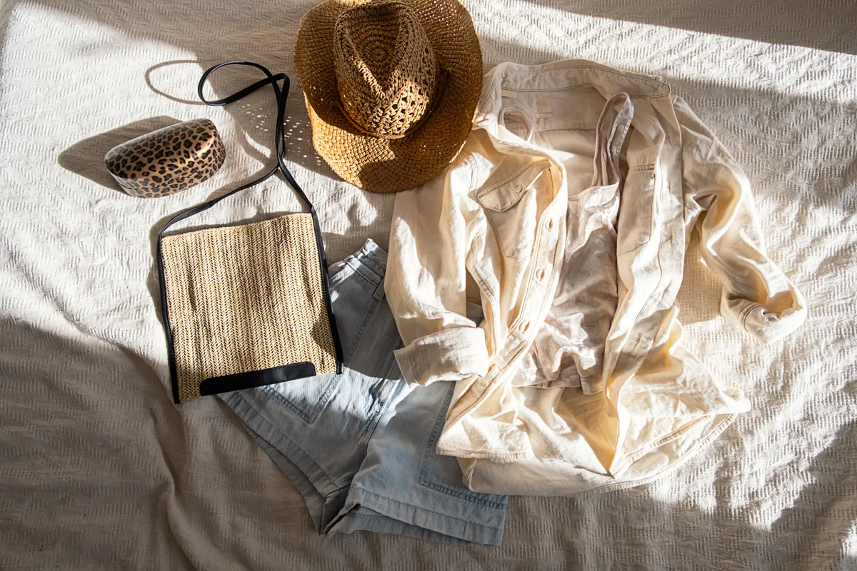 Sunlit flat lay of an ecru linen camp shirt, faded denim shorts, woven straw hat, and raffia crossbody bag on a textured bedspread