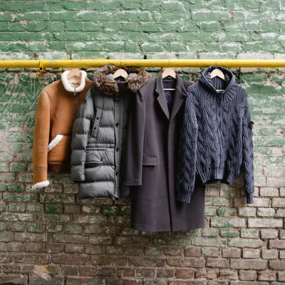 Charcoal wool city overcoat hanging from a yellow rail against a weathered green brick wall, photographed with its cold-weather companions — a tan shearling jacket, grey quilted parka, and slate cable-knit zip-up
