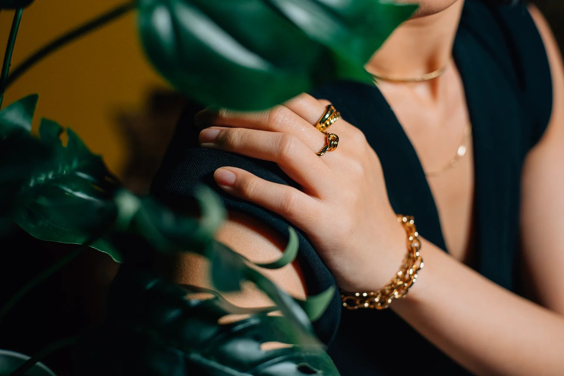 Model in a black dress wearing layered Aurelia gold — sculptural rings, a curb-chain bracelet and fine necklaces — among deep green leaves