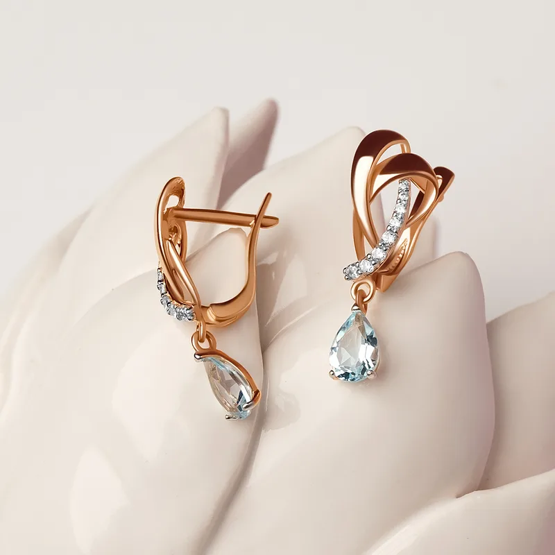Mar drop earrings in rose gold, pear-cut aquamarines hanging from pavé crossover hoops, staged on white sculpted petals