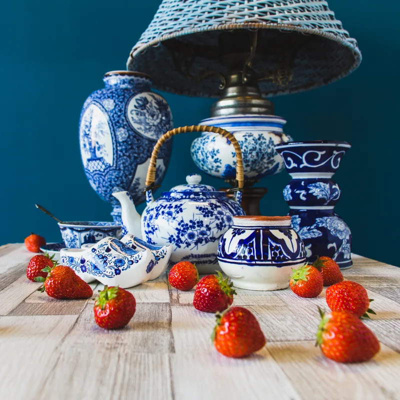 Blue-and-white patterned ceramics — a teapot, vases, and a wicker-shaded lamp — gathered on a pale wooden table with scattered strawberries