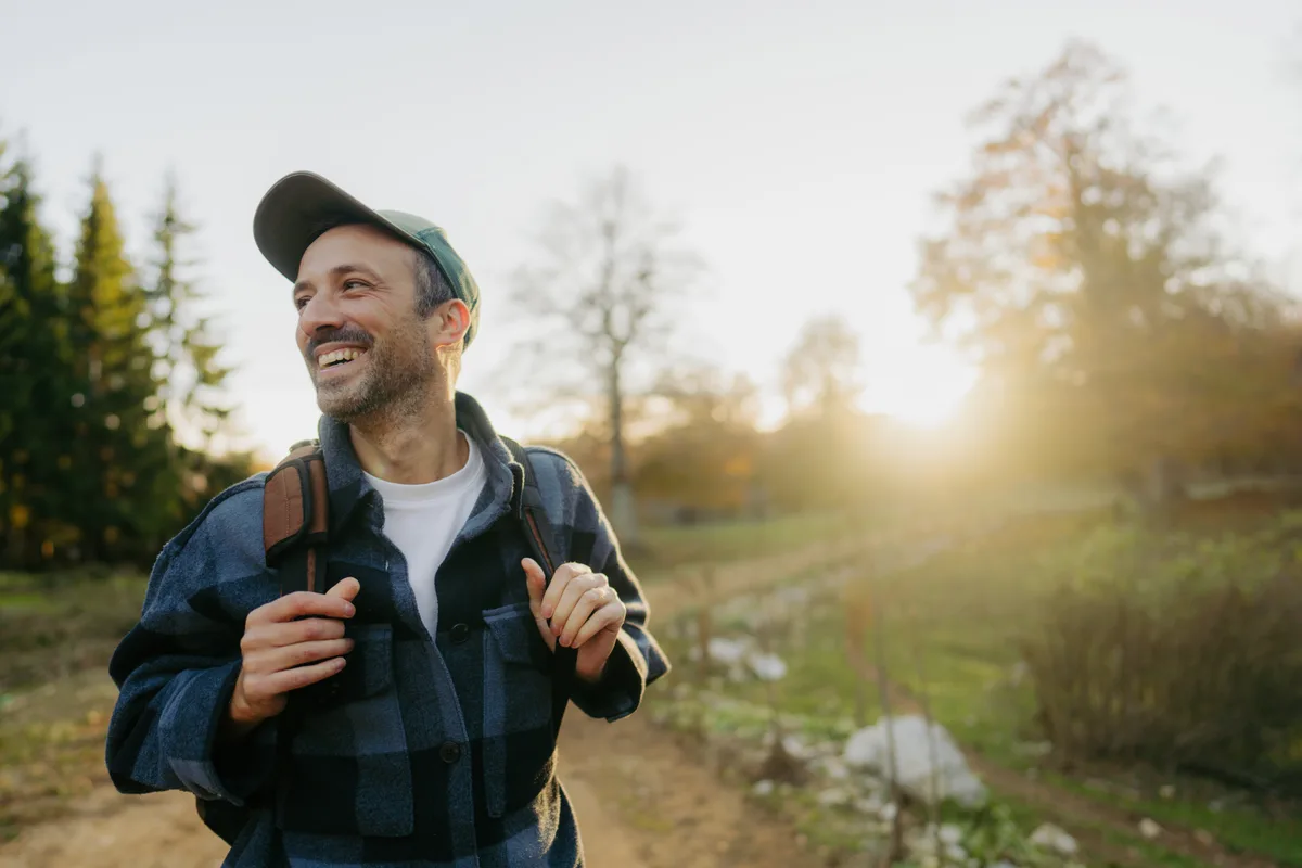Hiker in a plaid shirt grinning on a sunlit trail at golden hour, thumbs hooked under his backpack straps