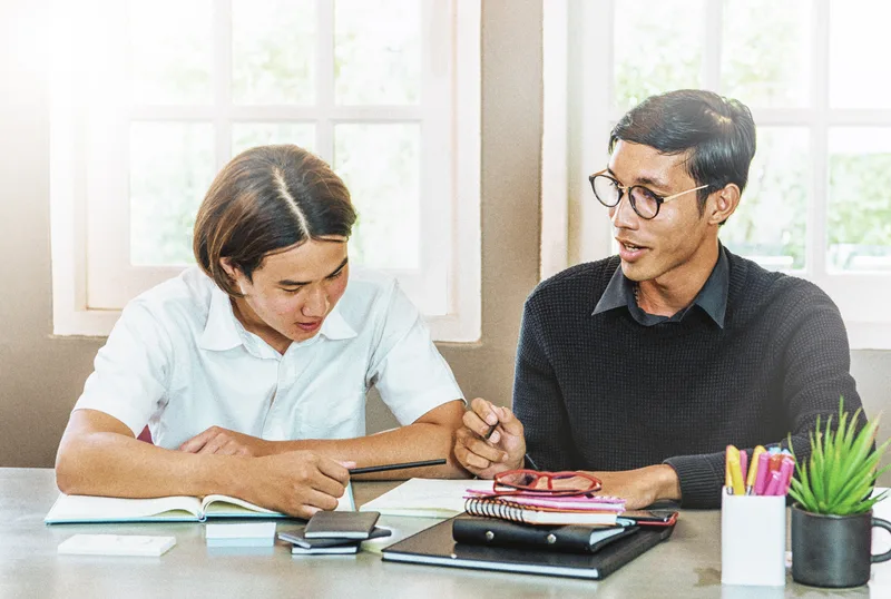A tutor explaining a concept to a teenage student across a study table stacked with notebooks
