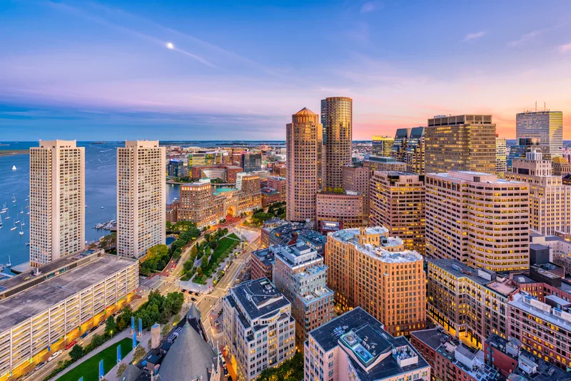 Aerial view of Boston's financial district and harbor at sunset, sailboats dotting the water
