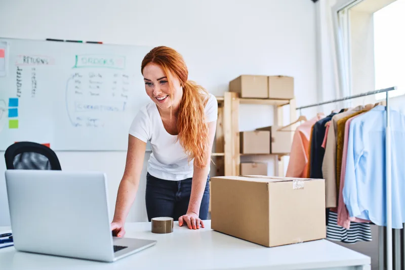 Online store owner checking orders on a laptop beside packed shipping boxes