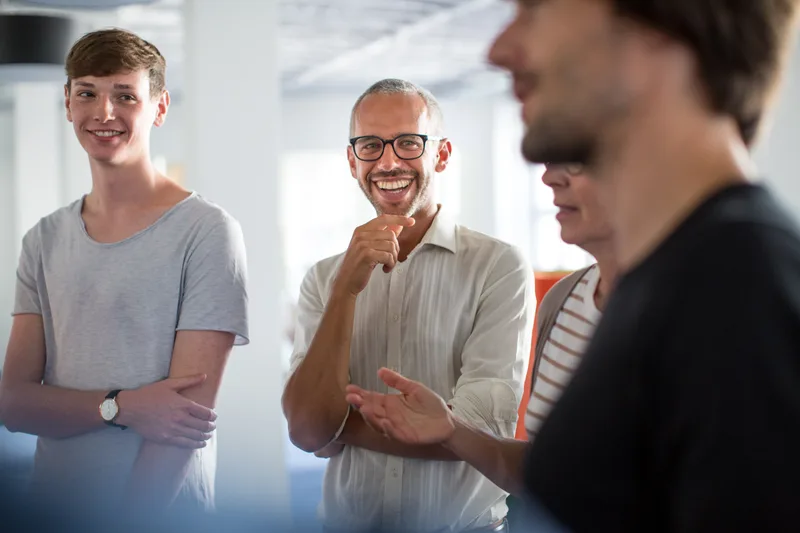 Agency team members laughing together during a relaxed stand-up meeting in a bright office