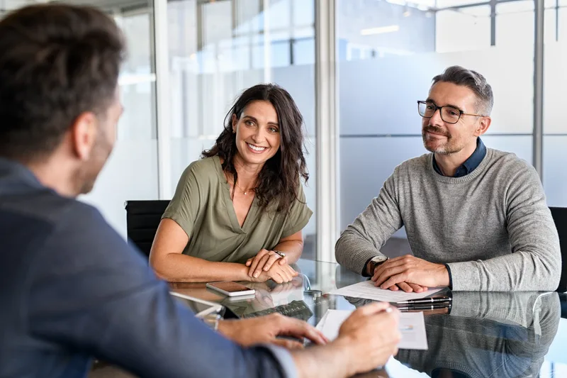 A smiling couple sitting across the desk from their insurance agent, policy paperwork spread out between them