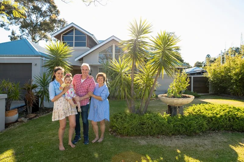 Three generations of a family — a mother holding her toddler, father, and grandmother — standing together on the sunny front lawn of their two-storey home