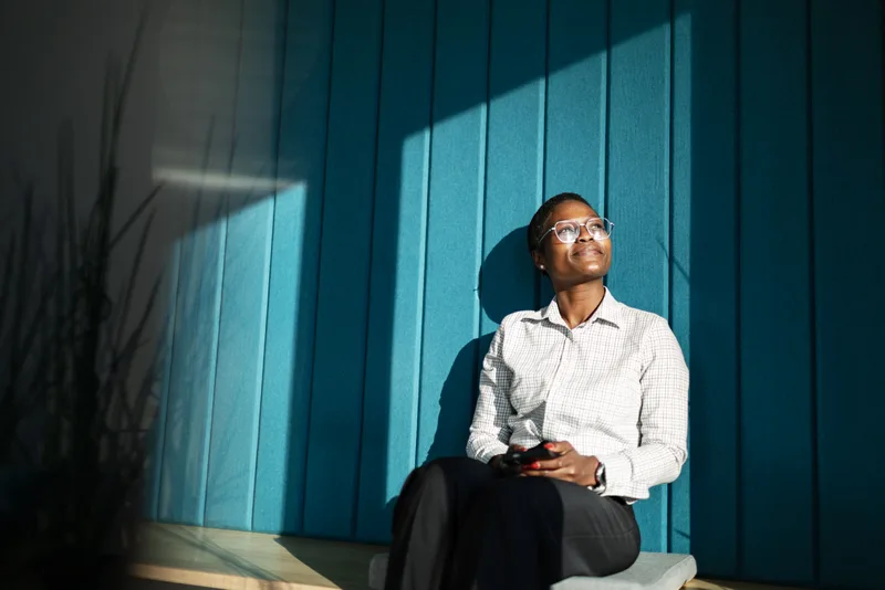 Woman in glasses and a checked shirt seated against a deep teal wall, lit by a hard diagonal shaft of afternoon sun