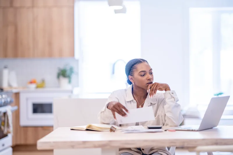 A woman at her kitchen table pausing over an opened bill, laptop and notebook in front of her
