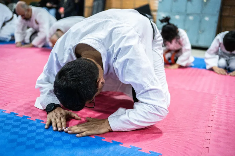Student in a white gi bowing deeply on interlocking mats while younger classmates kneel behind