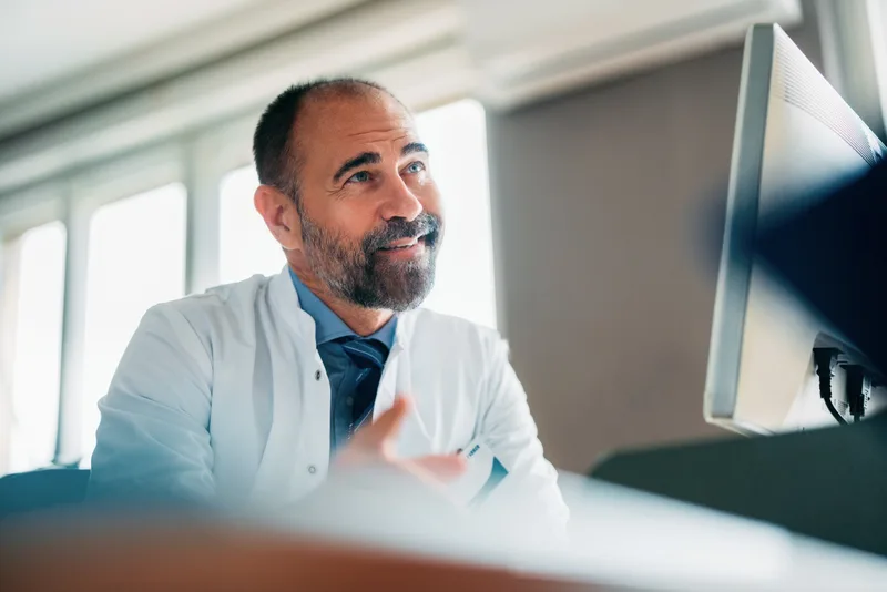 Dr. Peter Lindqvist in a white coat smiling at a desktop monitor in a bright office