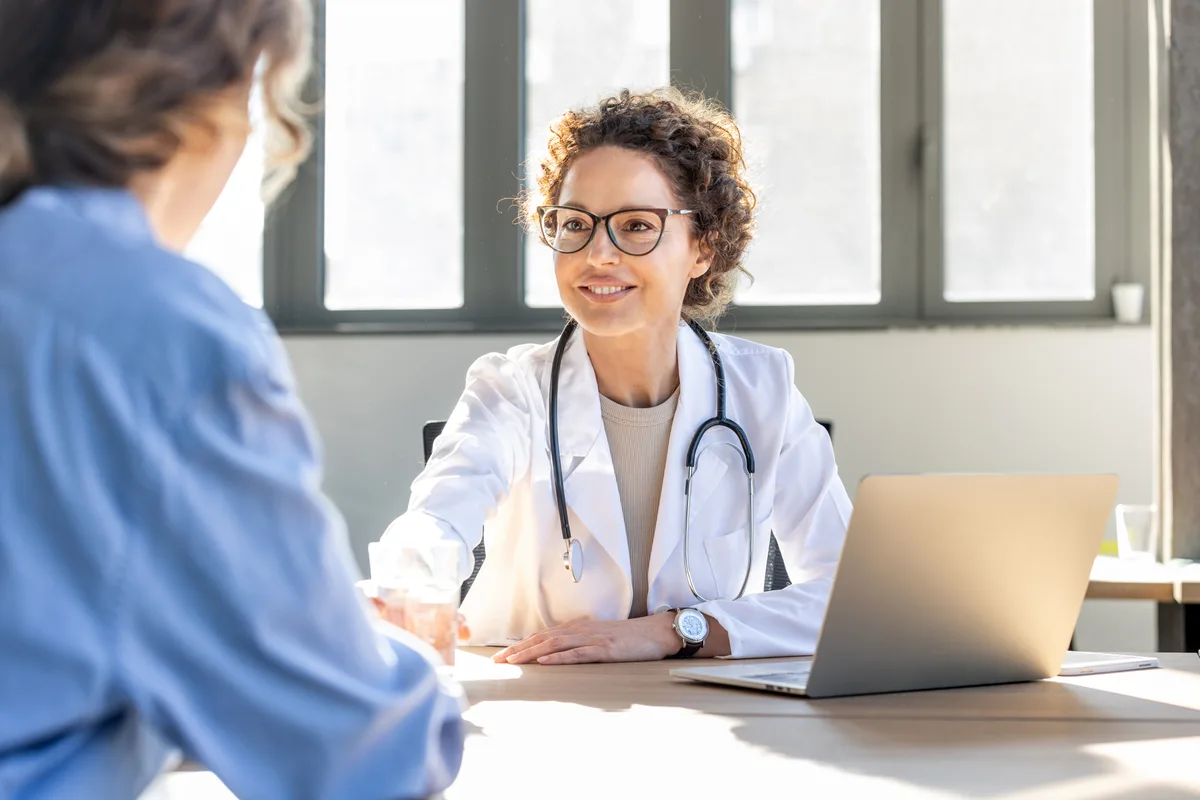 A physician in a white coat listening attentively to a member across a sunlit desk