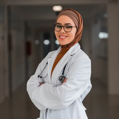 Dr. Amira Hassan, a physician wearing a headscarf, glasses, and a white coat, standing in a clinic corridor