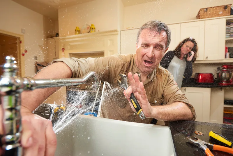 Homeowner shielding himself from a kitchen sink pipe spraying water while his partner calls for help