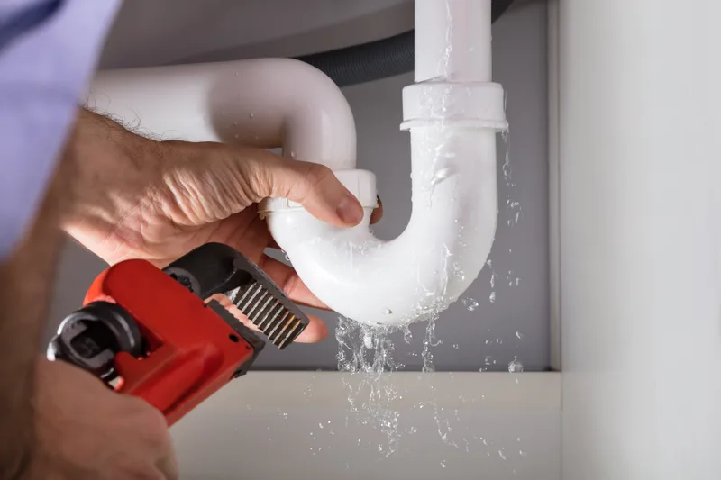Plumber loosening a leaking white sink trap with a pipe wrench as water drips out
