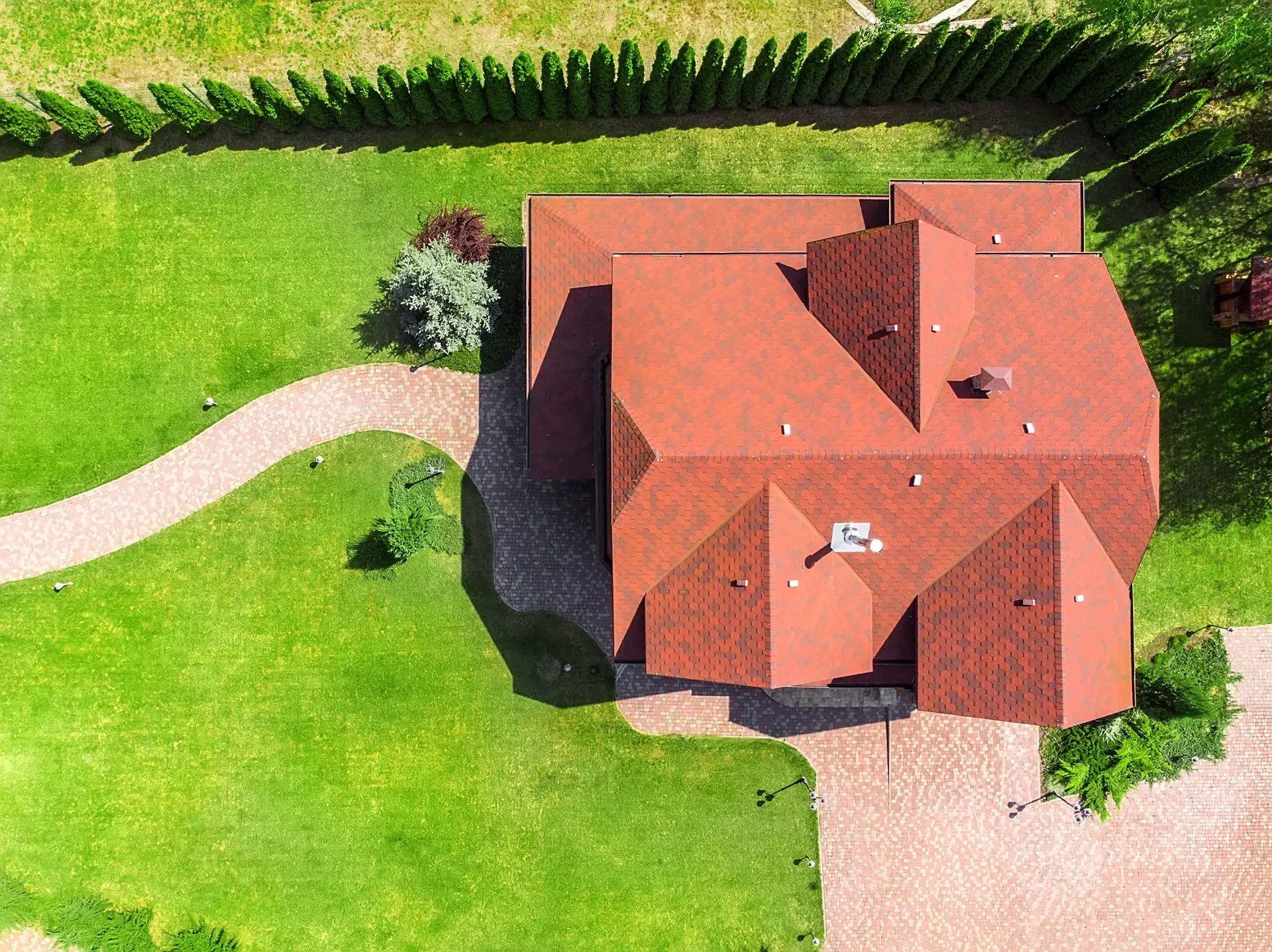 Aerial view of a red-roofed home with a curving paver path through deep lawn, edged by a tall cypress hedge