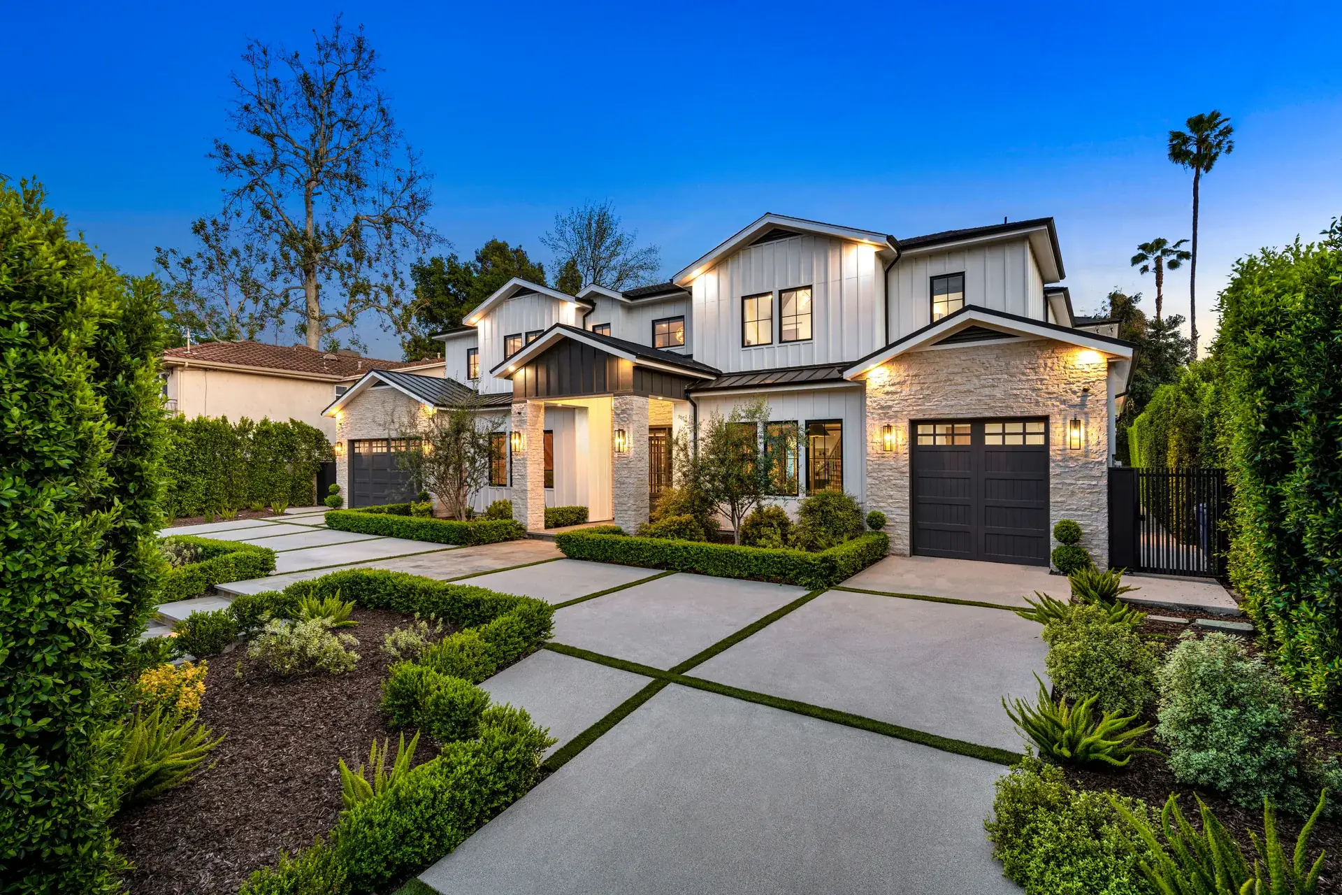 Modern farmhouse at dusk with warm uplighting, clipped hedge frames and a concrete-panel auto court with turf joints