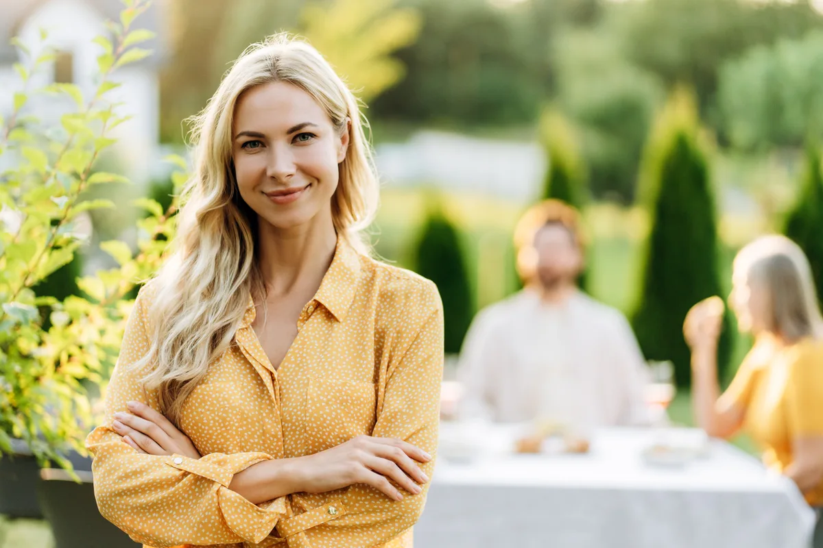 A woman smiles with her arms folded at a relaxed multigenerational dinner in a sunlit garden