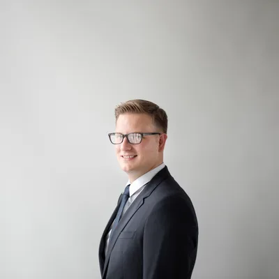Tom Lindqvist, a smiling young man with glasses in a charcoal suit, photographed against a soft grey background