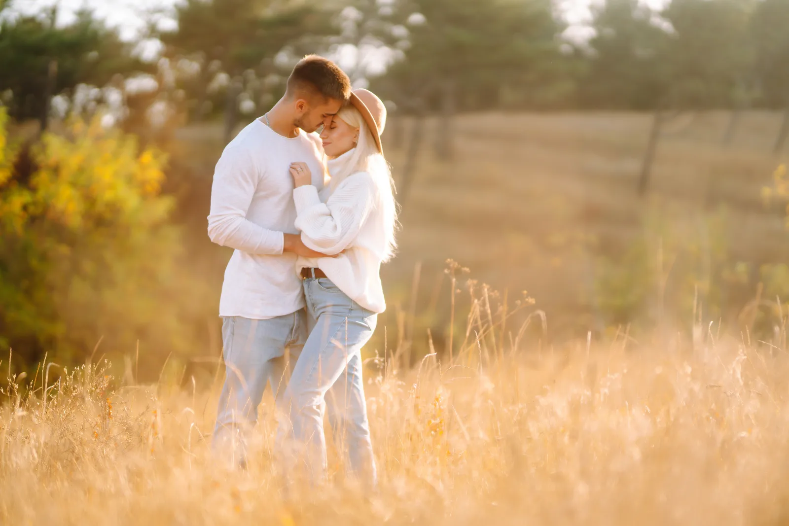 Couple in white sweaters embracing forehead-to-forehead in a golden backlit field at sunset