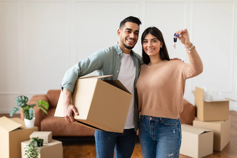 A young couple holding up the keys to their first home, moving boxes stacked behind them