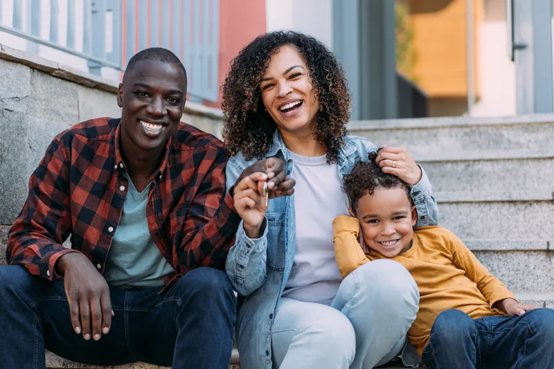 Parents and their young son sitting on the front steps of their new home, holding the house keys