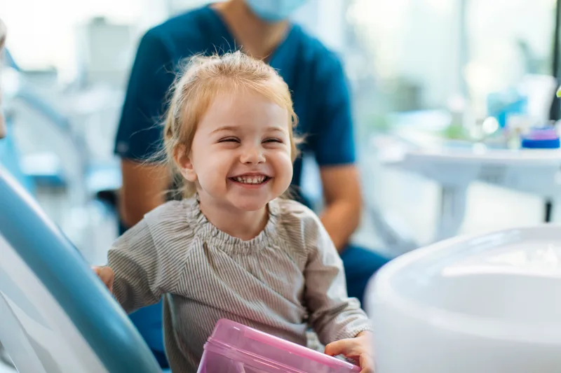 A toddler laughing in the dental chair, clutching her pink prize box, clinician smiling behind her