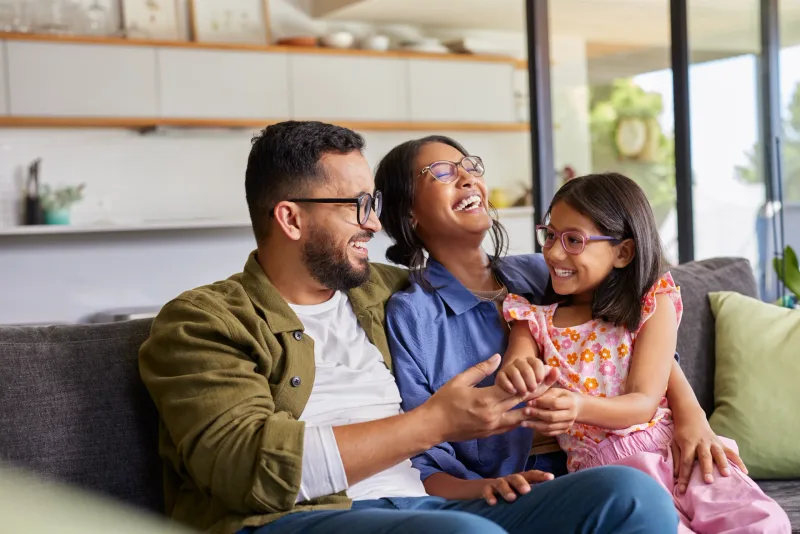 Father, mother, and daughter in glasses laughing together on a grey couch at home