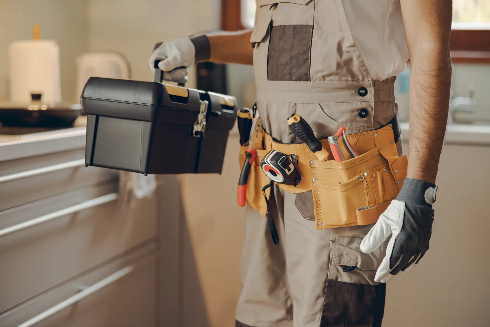 Handyman in a loaded tool belt carrying his toolbox into a client's kitchen for a repair visit