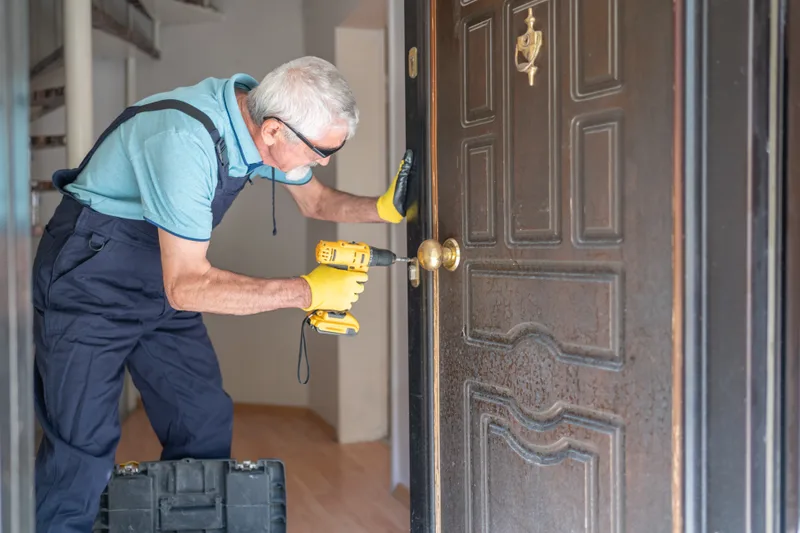Locksmith in work overalls fitting a new deadbolt into a dark wooden front door with a cordless drill