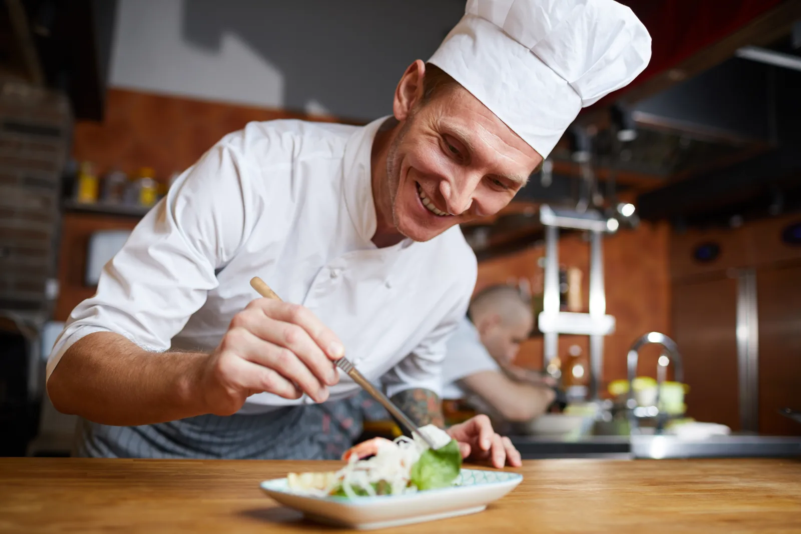 Chef plating an entrée at the pass during dinner service in a busy restaurant kitchen