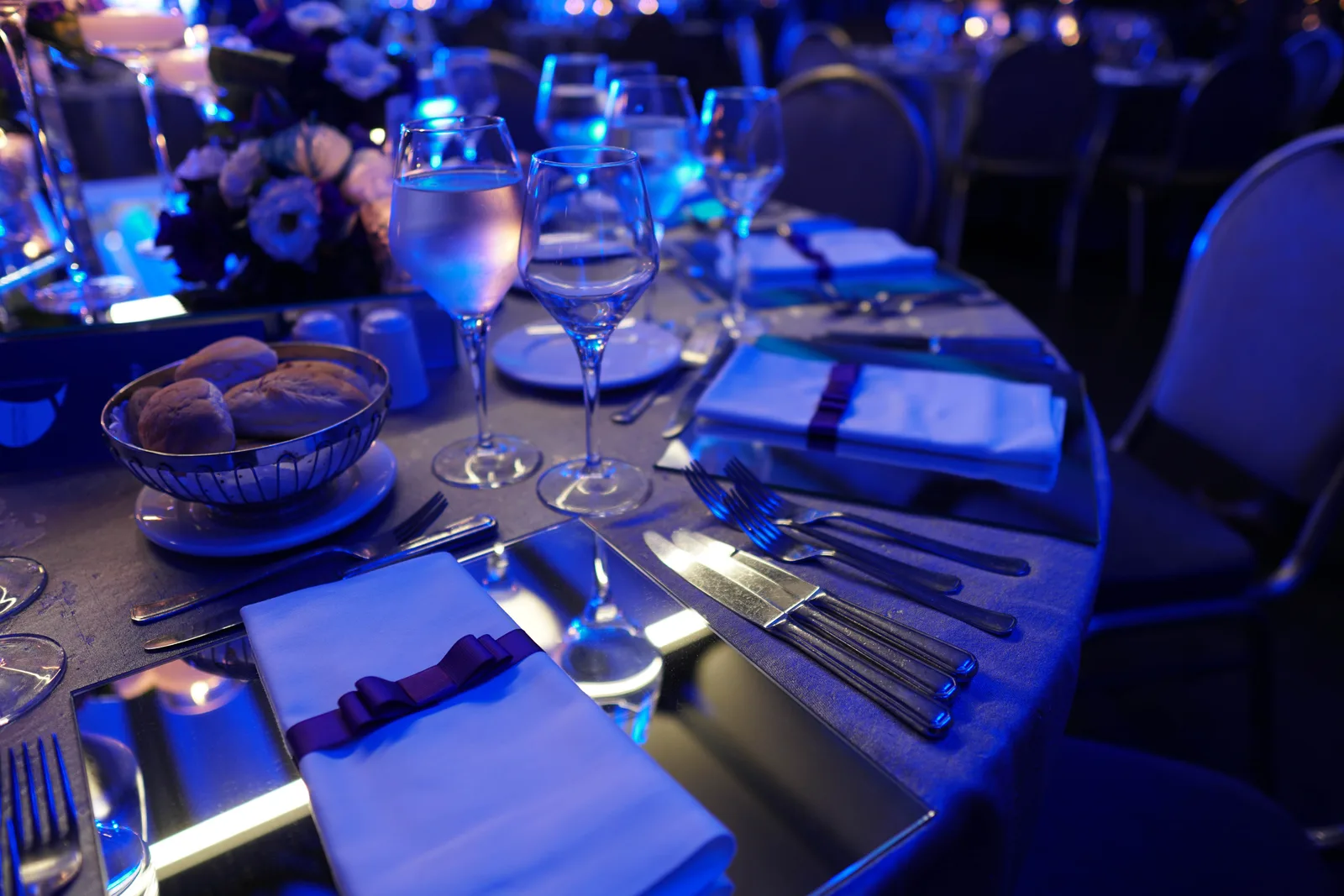 Reception table at a wedding venue set with glassware, ribboned napkins, and florals under blue uplighting