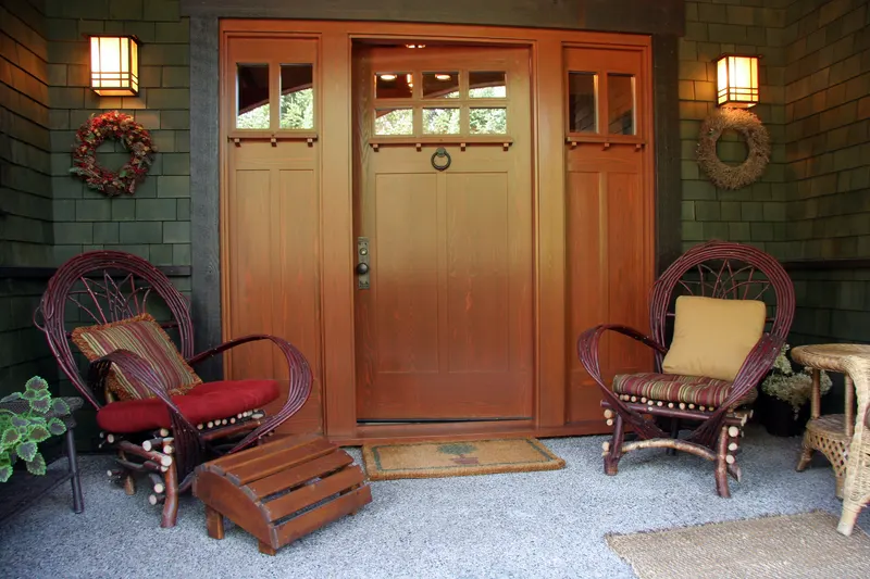 Deep craftsman bungalow porch with green shingle siding, a wide wood front door with sidelights and twig rocking chairs