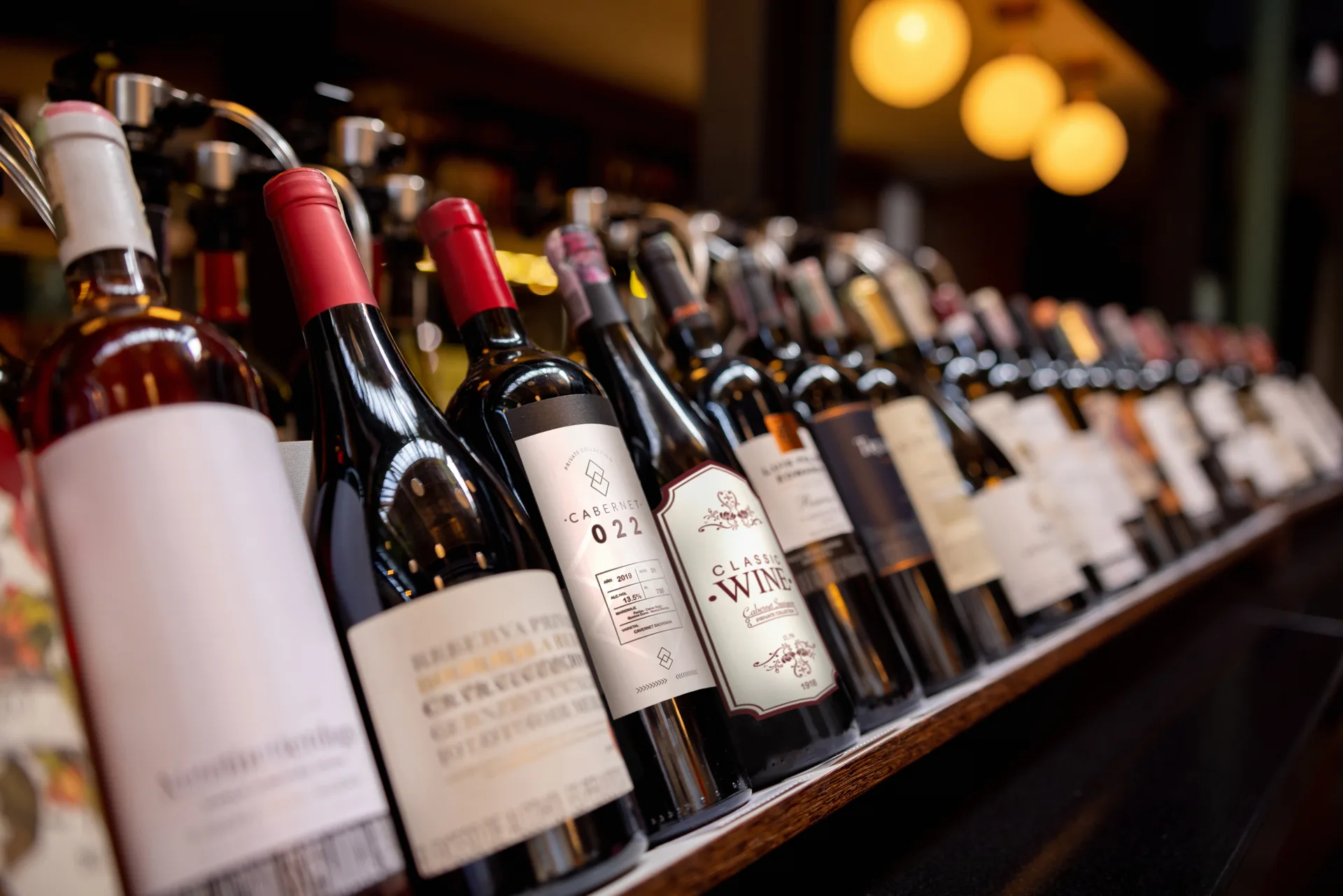 A long row of wine bottles standing shoulder to shoulder on the anteroom's back bar, warm lamplight behind them