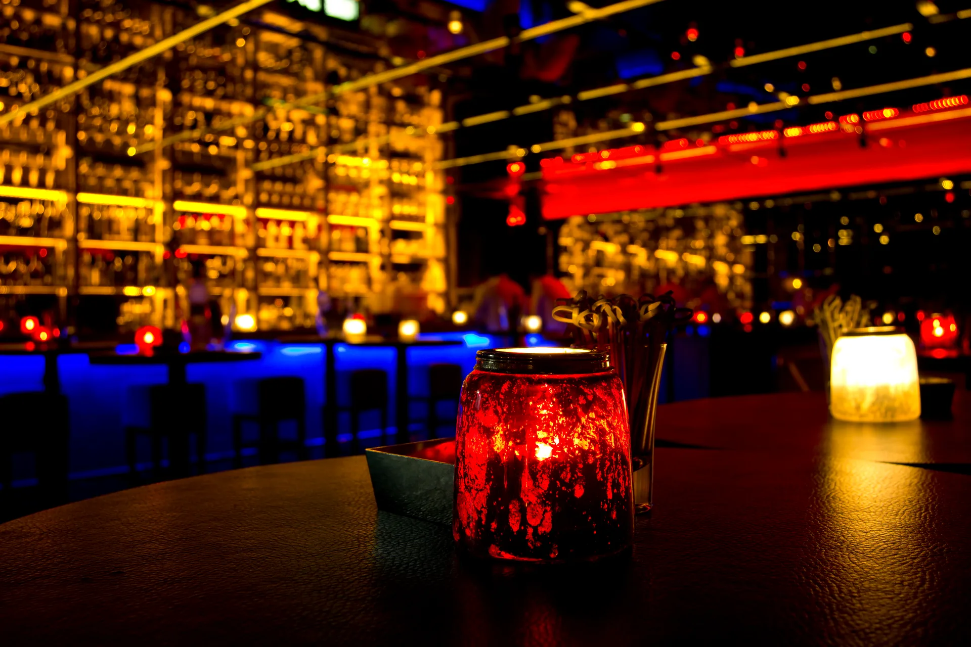 The anteroom bar at night — a candle flickering in a red glass jar on the counter, backlit bottle shelves glowing behind