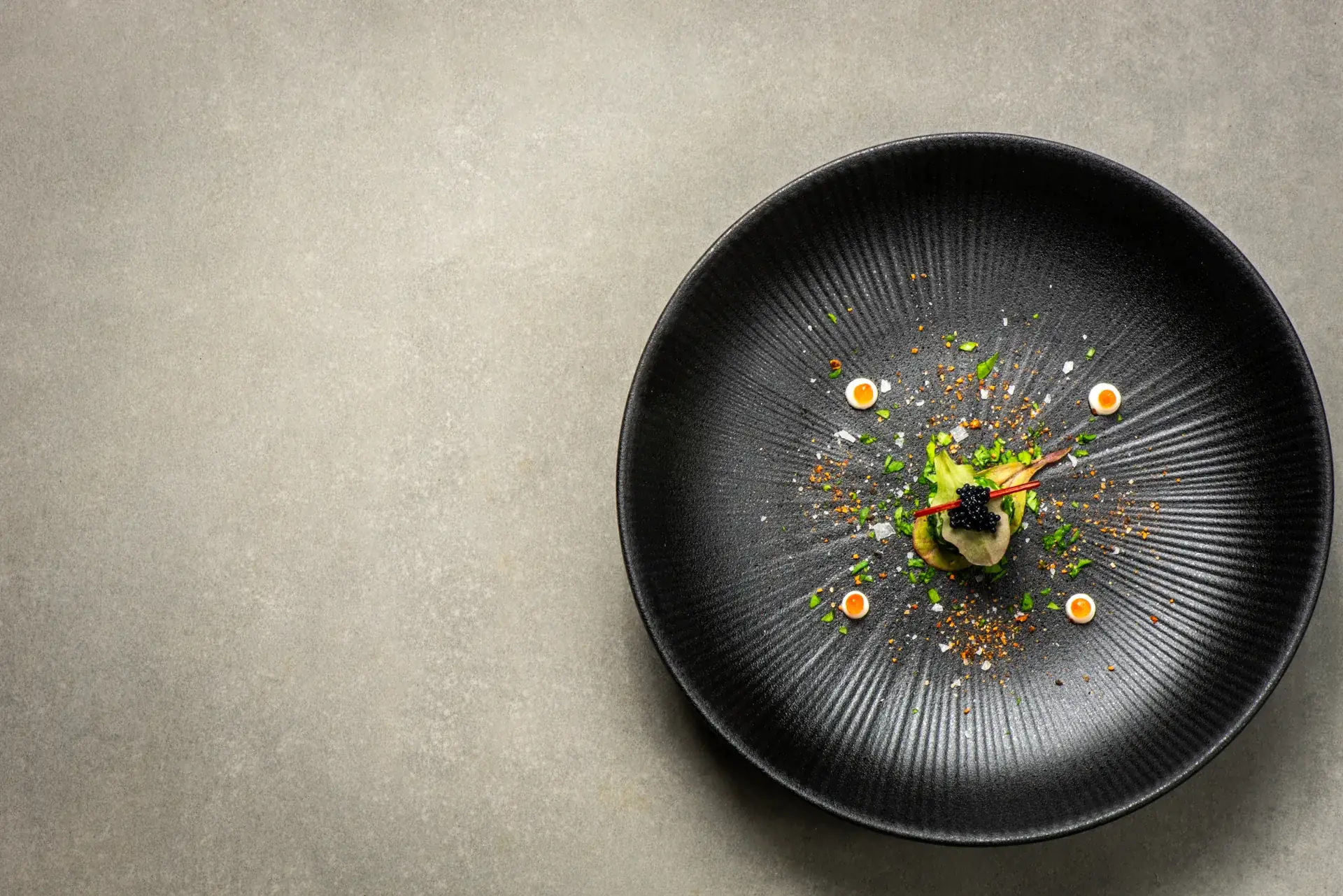 Overhead view of a single composed bite — avocado, caviar, and herbs — centered on a black ridged plate against gray stone