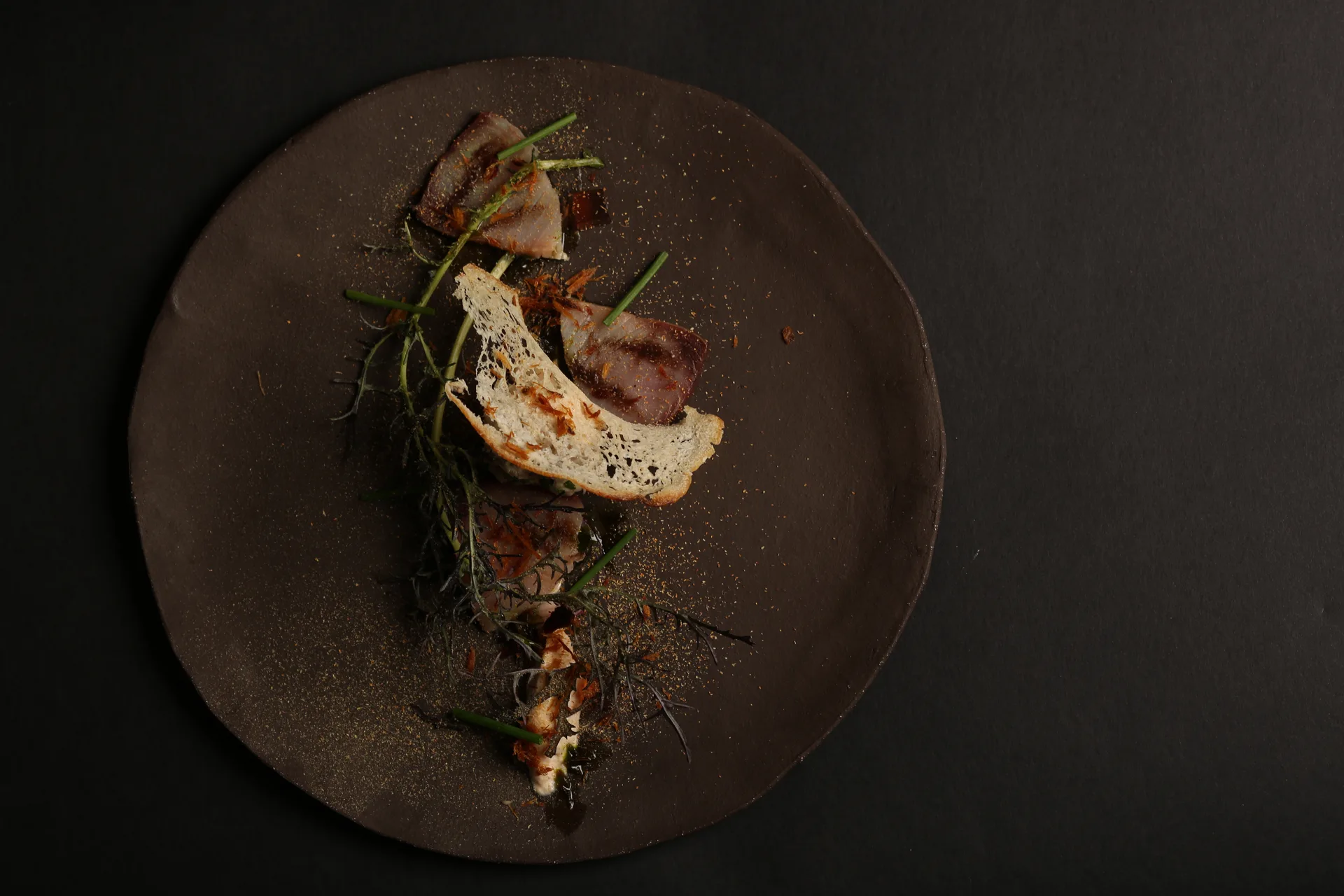 A foraged course arranged down the center of a dark ceramic plate — cured fish, crisp bread, and wild greens against near-black